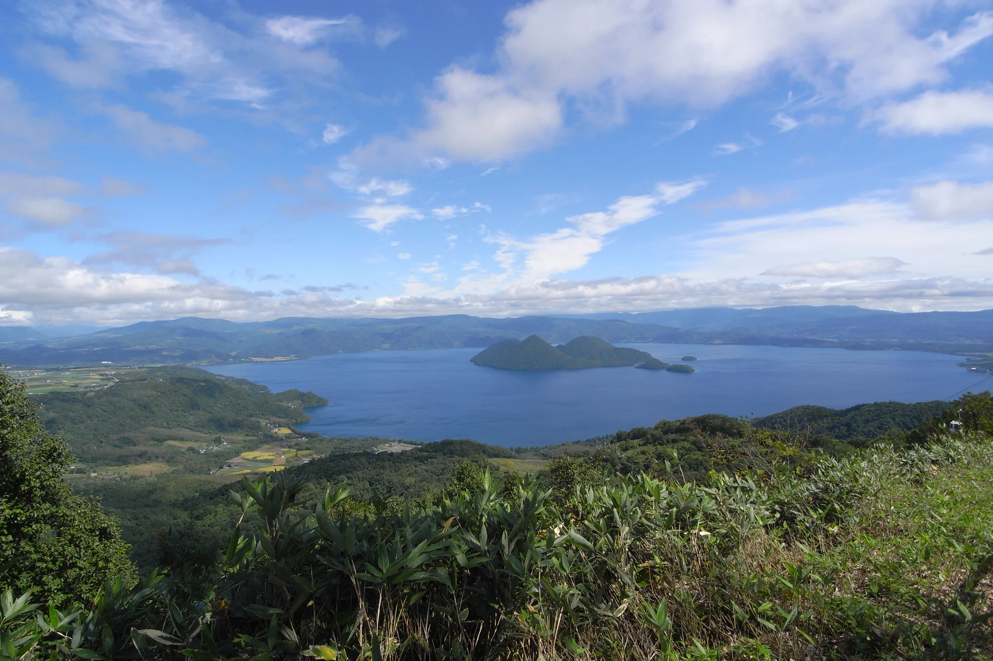 洞爺湖溫泉街全景，從湖畔步道望向沿岸飯店群與中島，遠處有珠山輪廓清晰