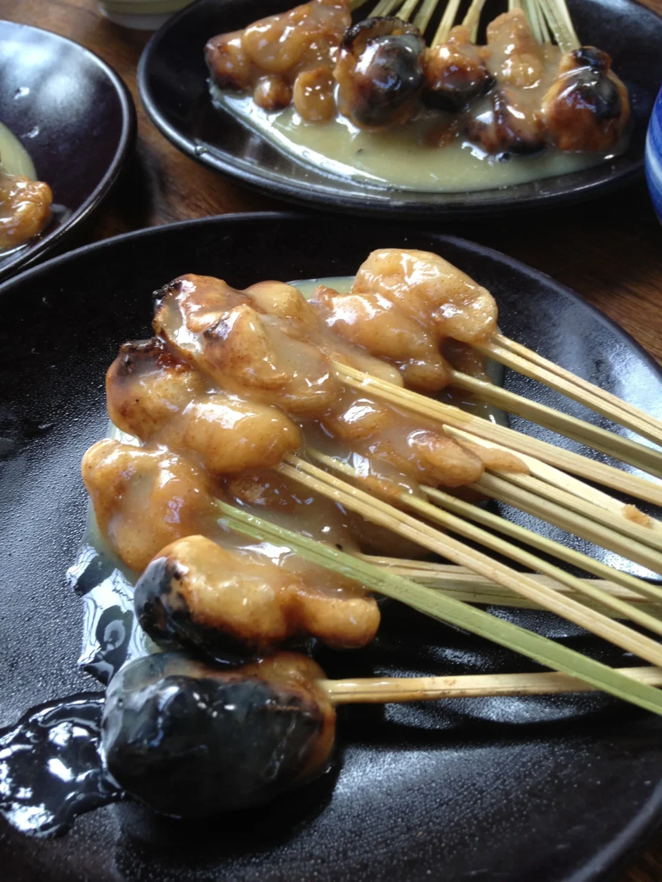 黑色陶碗盛装日式烤鸡串,竹签穿肉球配青葱,覆照烧酱汁,室内灯光。