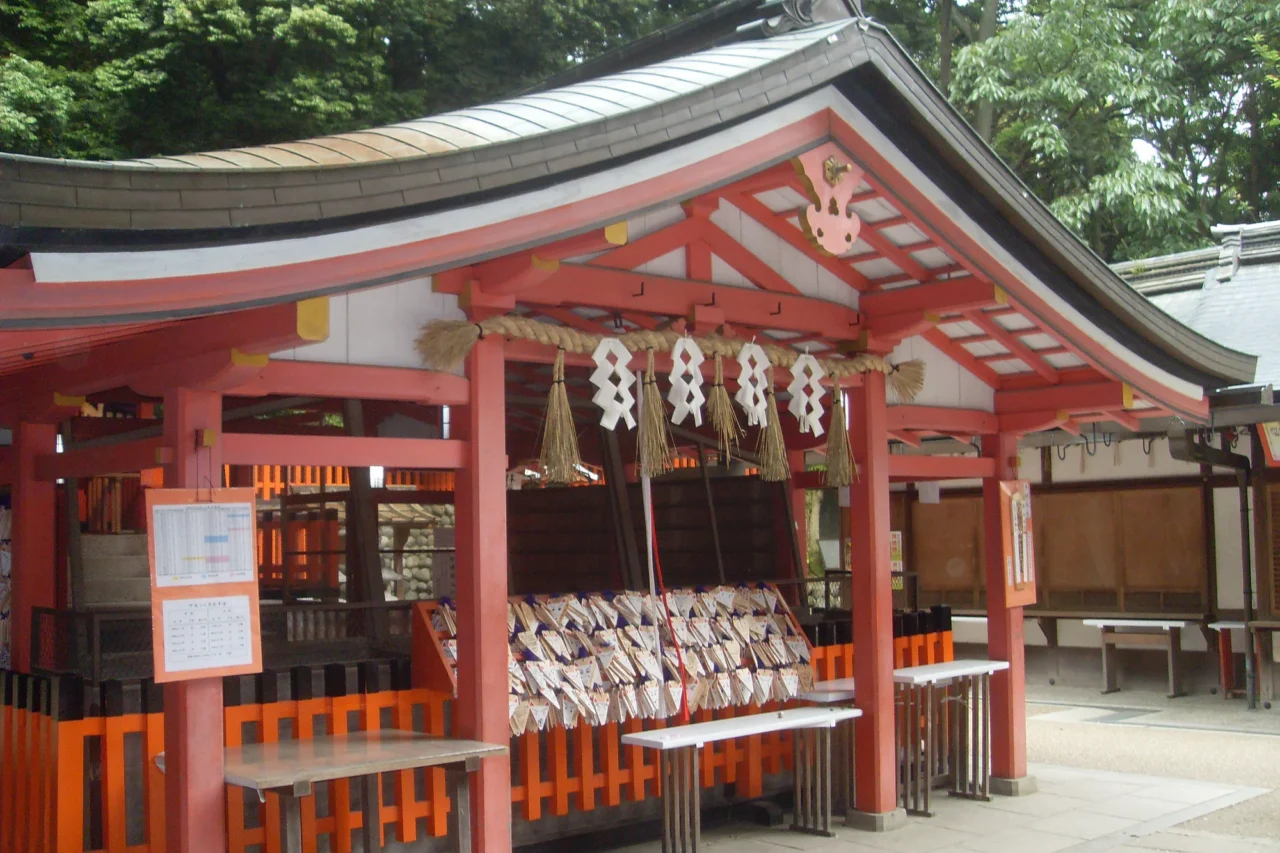 紅色木製神社建物，弧形黑屋頂、懸掛白色繪馬球、橘紅欄杆、綠樹環繞、白晝照射。