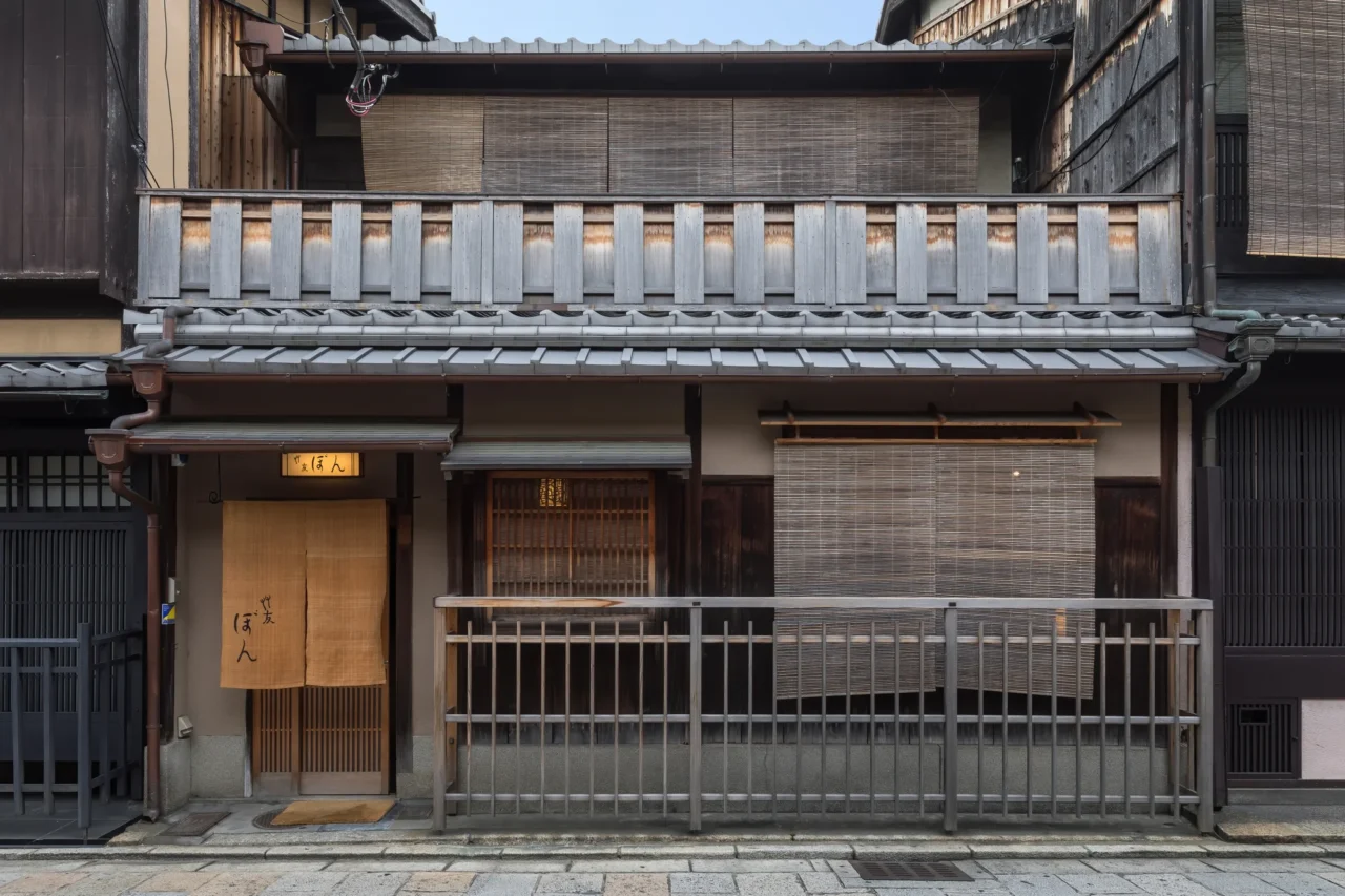 日本木造建筑，入口黄色招牌標記「保八」，竹格欄杆與深色木框架、灰瓦屋頂，白天石板路地面。