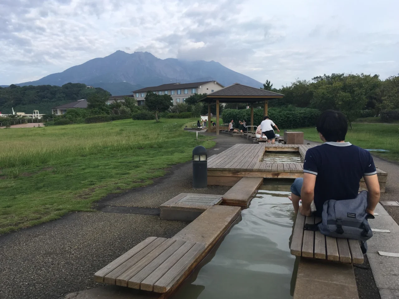 男性身穿黑衣坐在木制长凳上，远处绿草坡上矗立火山山脉，晴朗午后天空飘白云。