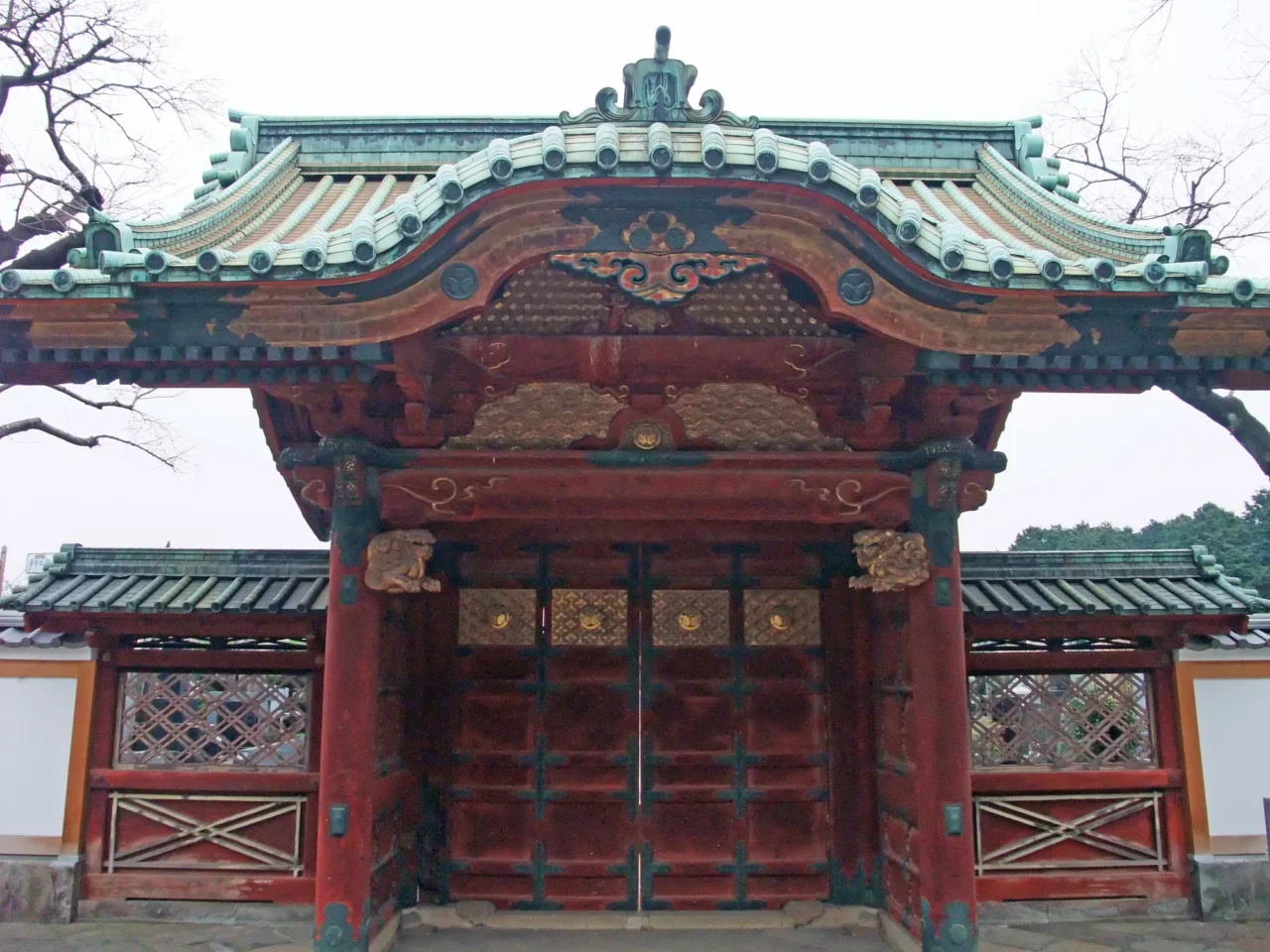 冬季陰天下，傳統廟宇的紅漆正殿，綠藍飛檐頂配金色裝飾，兩側對稱幾何牆，周圍光禿樹木。