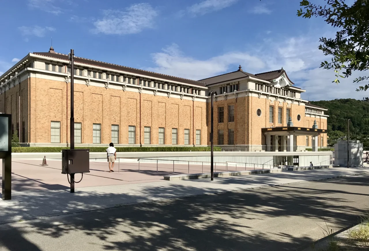 淺黃色古典磚石建築坐落寬敞廣場邊，晴朗藍天下樹影投映地面，多層窗戶整齊規則排列。
