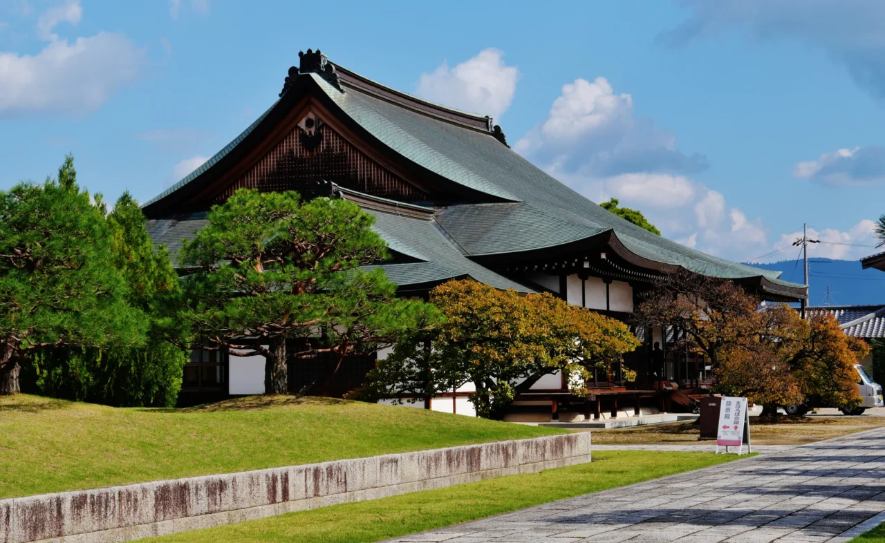 傳統日本寺廟建築配黑色瓦屋頂，周圍綠色庭園、白色石欄杆環繞，晴天藍天白雲背景。