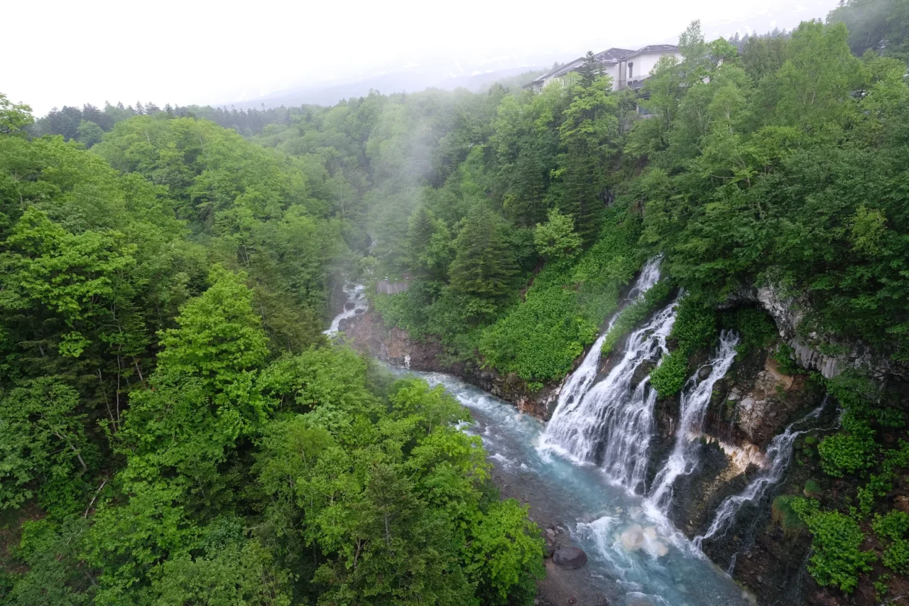 白色瀑布從岩壁傾瀉入青藍色溪流,兩岸密集綠林籠罩,陰霧中的山澗峽谷。