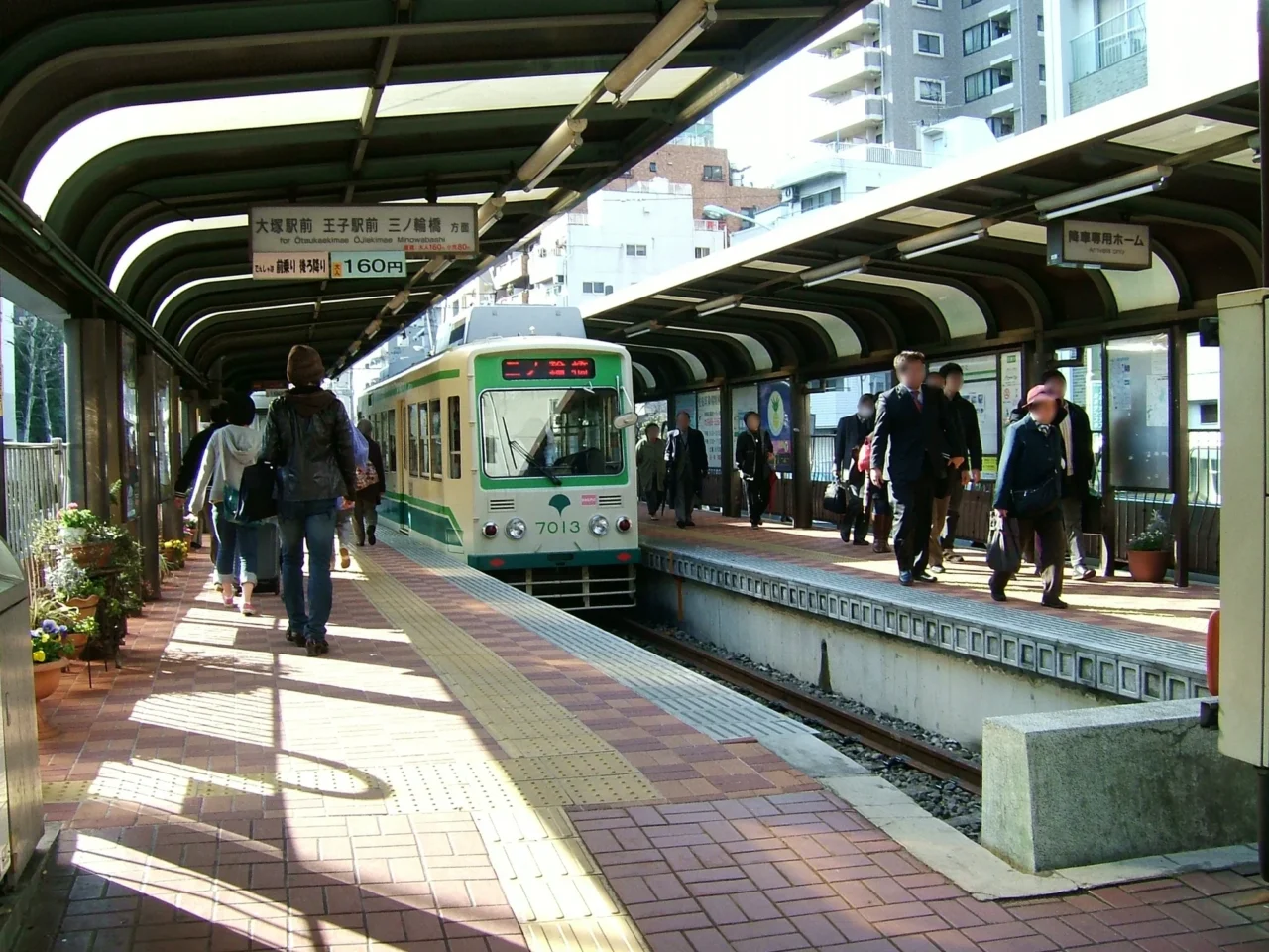 綠色火車靠站月台，旅客在有遮蓋棚的紅褐色月台候車上下，背景為現代住宅樓，日光充足。