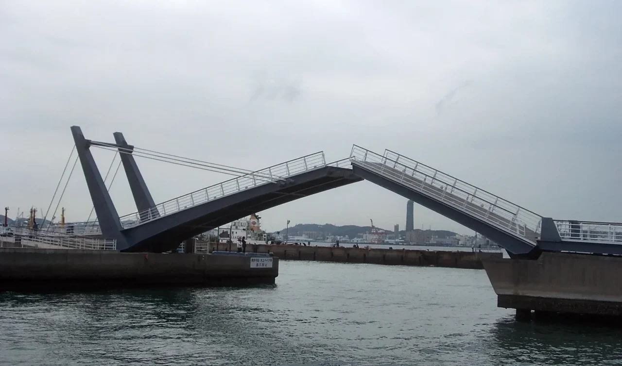 藍翼門司開合橋黑色橋身傾斜,港灣水面展開,背景工業建築羣,灰色天空。