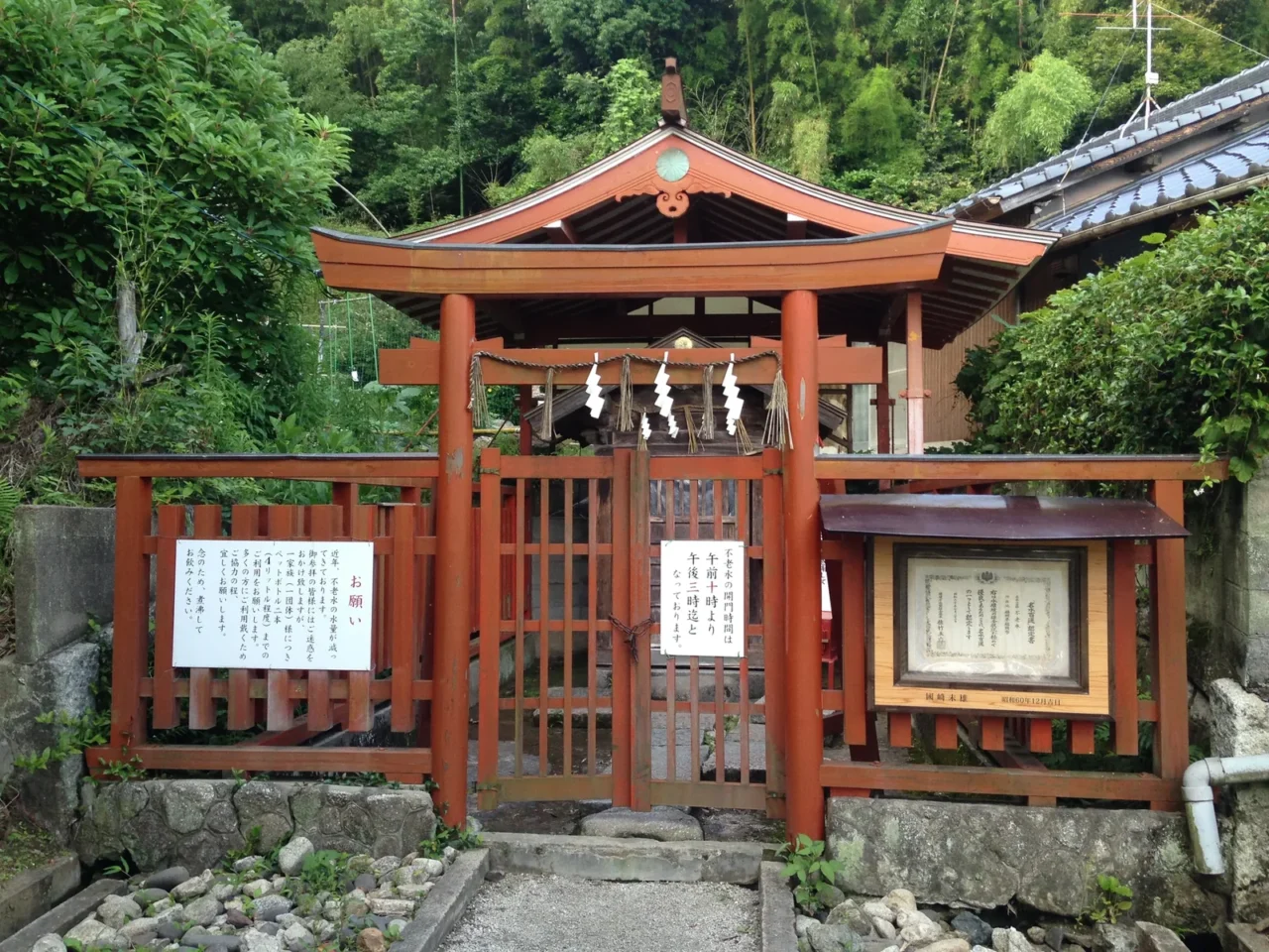 不老水木製神社入口，尖頂屋頂懸掛白色紙籤，綠樹密林環繞。