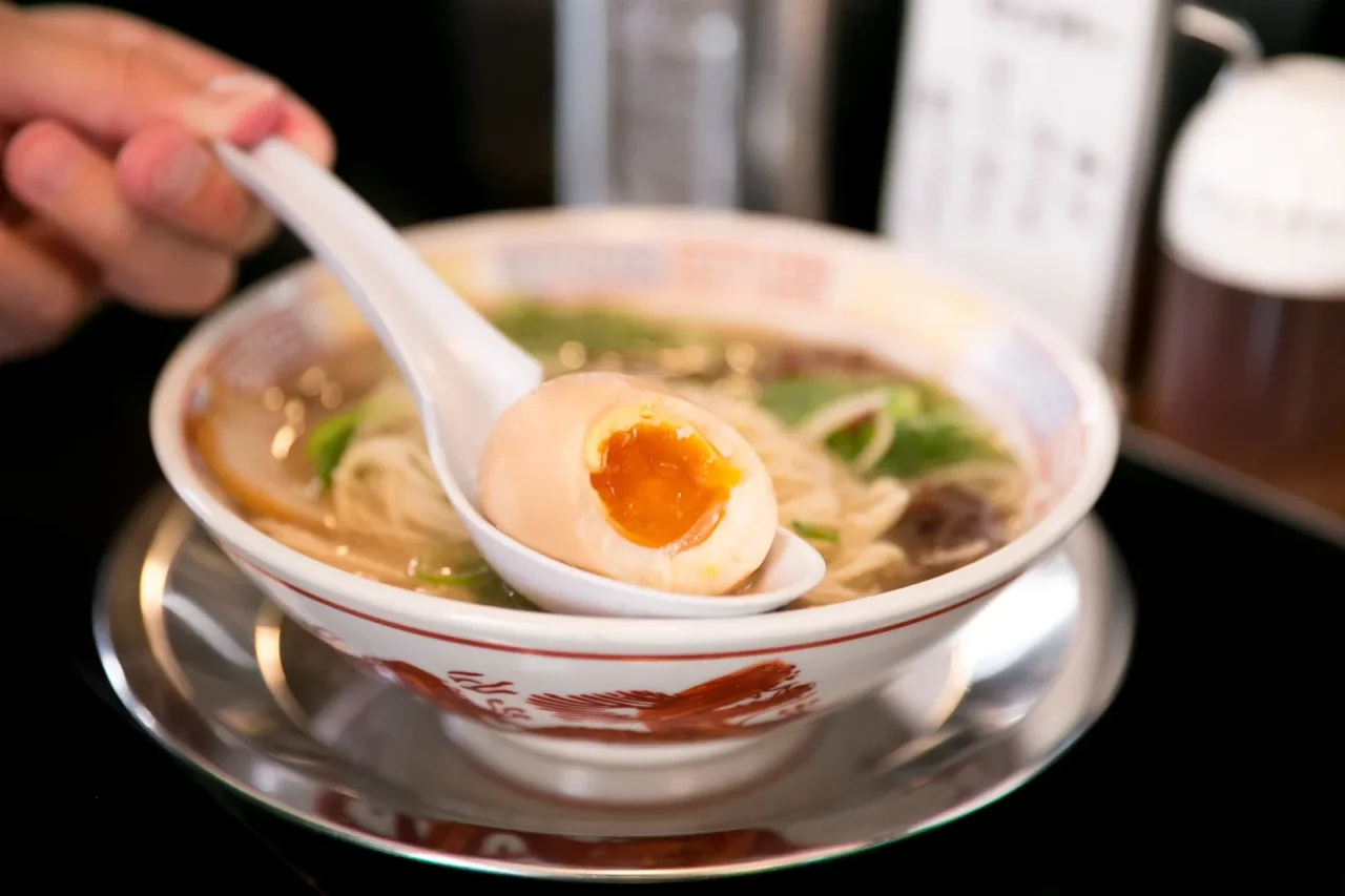 博多めん街道拉麵碗盛放湯麵與溏心蛋,灑青蔥碎,室內昏暗用餐環境。