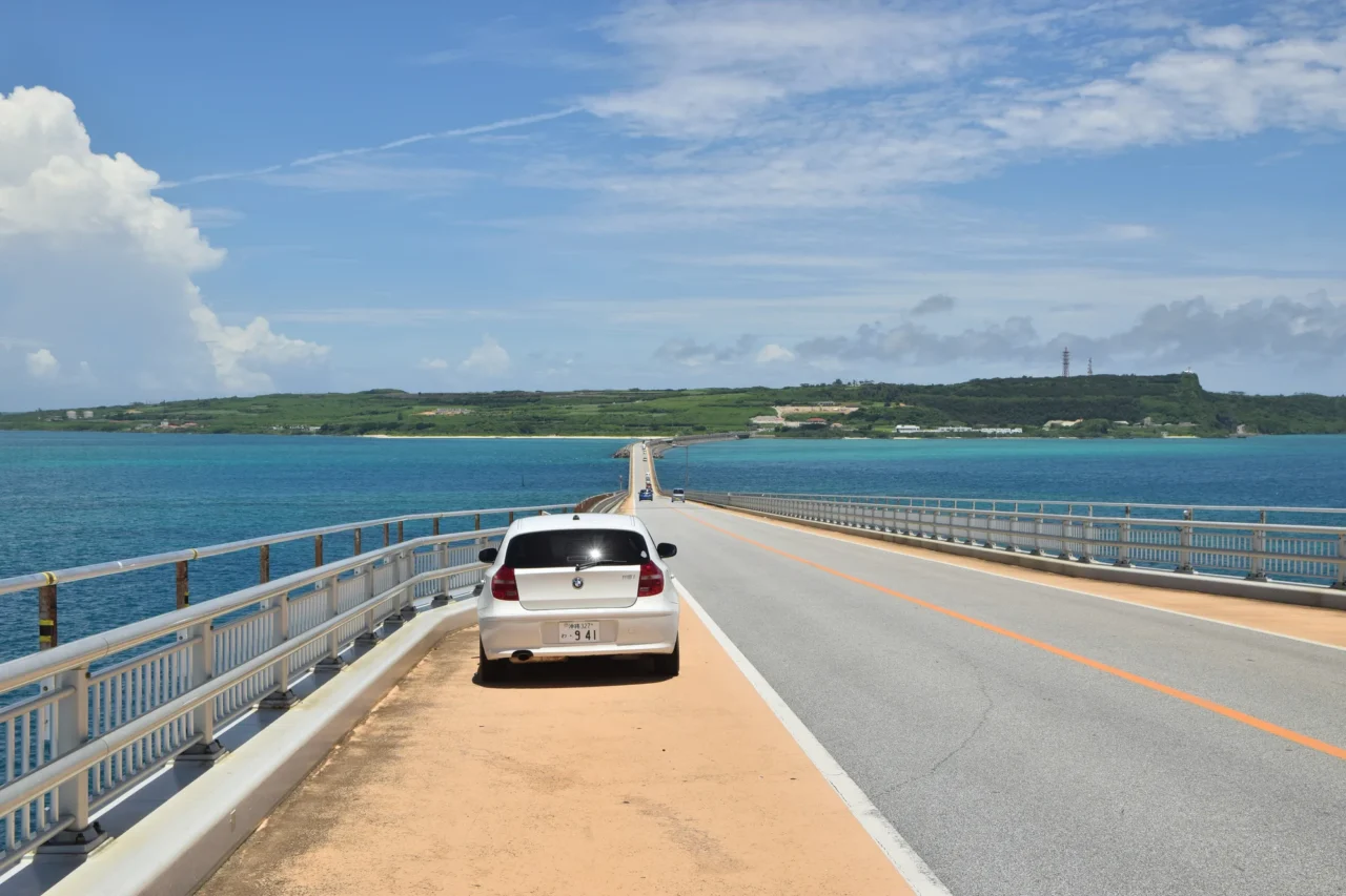 宮古島跨海大橋上白色轎車停靠沙土區,護欄與道路延伸向海對岸島嶼。