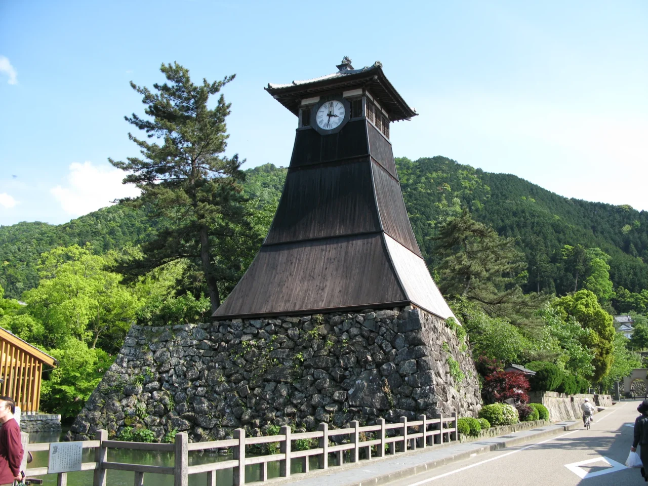 出石時鐘塔黑尖頂與石塊基座,白木柵欄環繞,山林中遊客漫步。