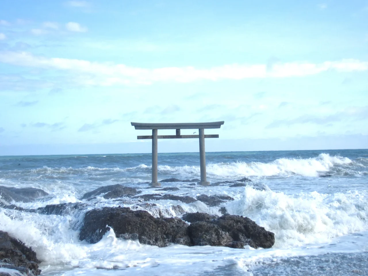 大洗海邊灰色日式鳥居,屹立黑色巖石之上,白浪衝擊,晴空背景。