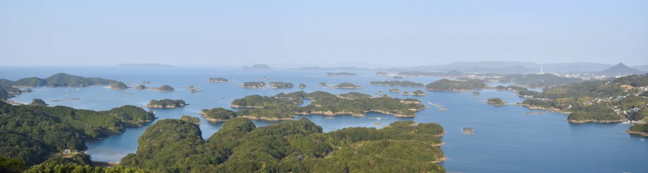 九十九島珍珠海洋遊覽區內海散布眾多綠島，陸地建築與山巒隱約可見，灰白天空映襯波光。