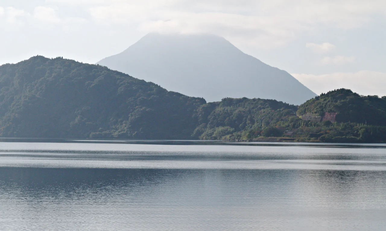 開聞岳與濃密松林相環，平靜湖水映照火山身影，陰天白雲籠罩全景。