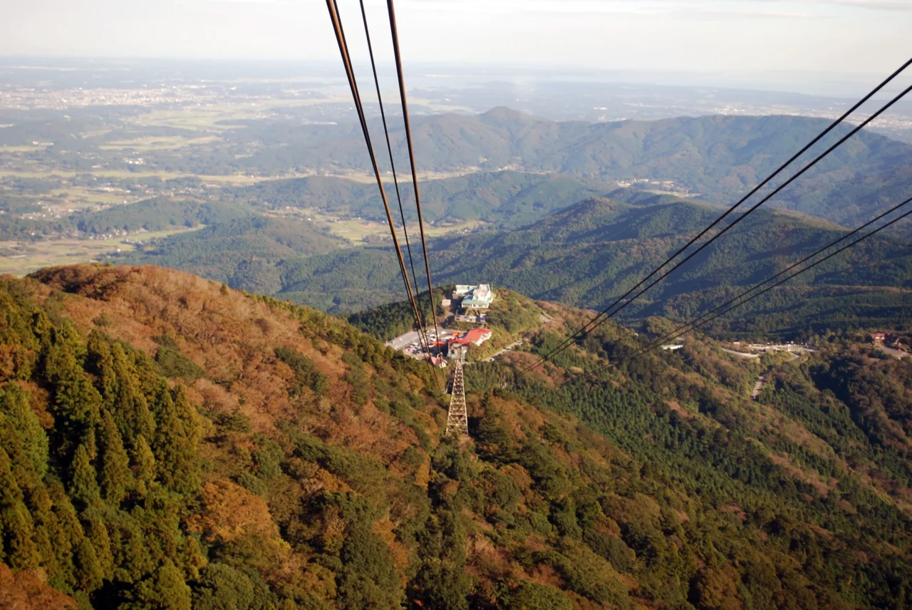築波宇宙中心山頂纜車穿過秋季紅褐樹林,俯視平原籠罩薄霧,晴朗日光照亮白色建築。