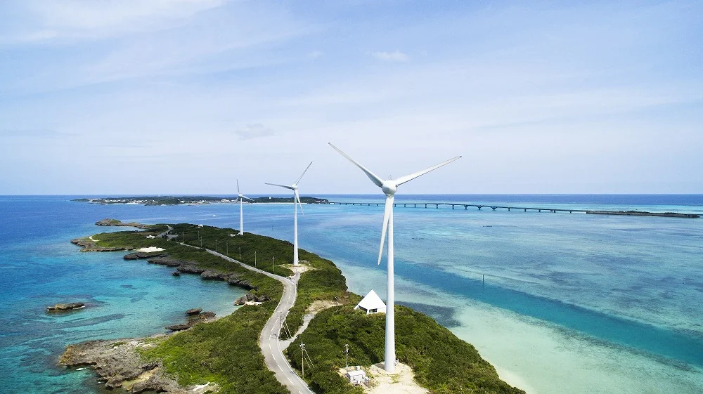 西平安名崎空中俯瞰,蜿蜒白色道路與風機點綴地形,遠方跨海大橋連接海岸,藍綠海水。