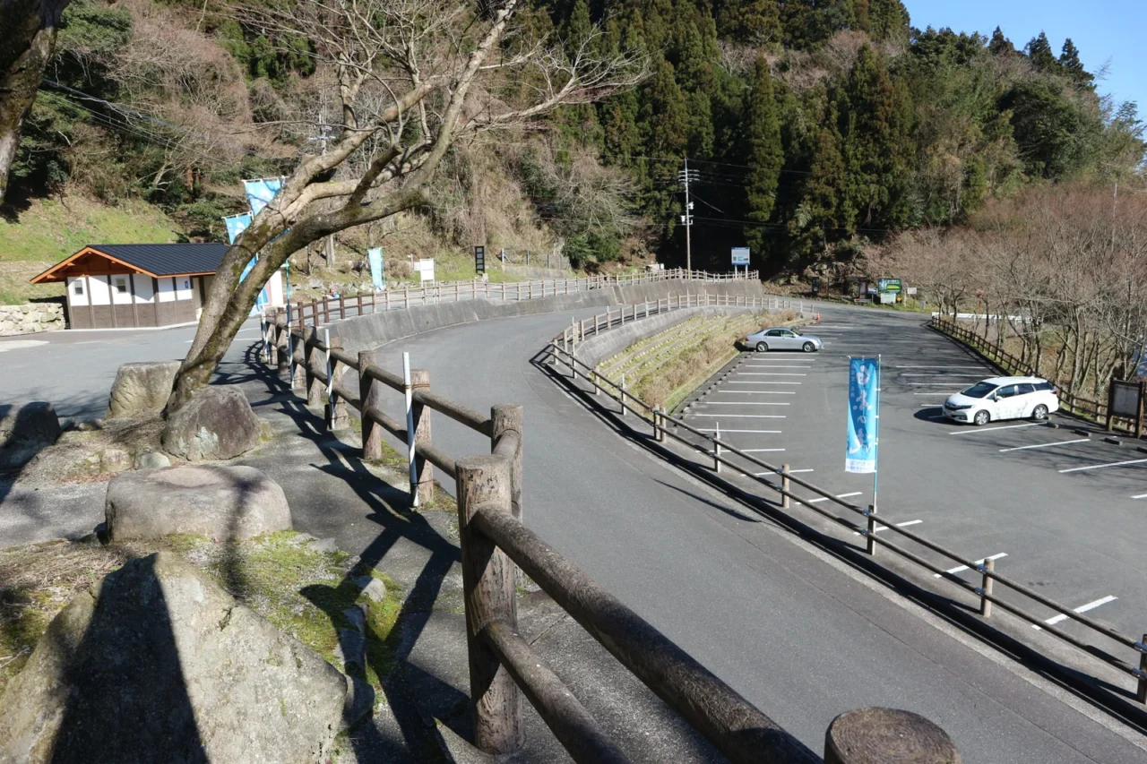 白糸瀑布大眾運輸停車場，古樸日式小屋旁停放汽車，大樹投影地面，背景為杉樹與晴空。