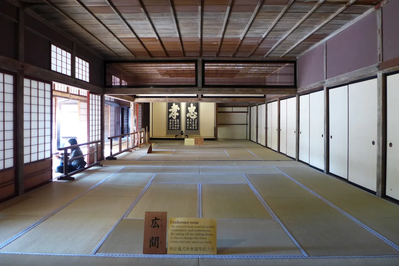 高山陣屋榻榻米廣間，訪客靠窗而坐，懸掛日文書法，白拉門與格子窗投入自然光。